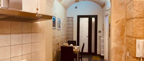 A narrow kitchen with a stone archway, a gas stove, a dining table with chairs, and a door leading to another room.