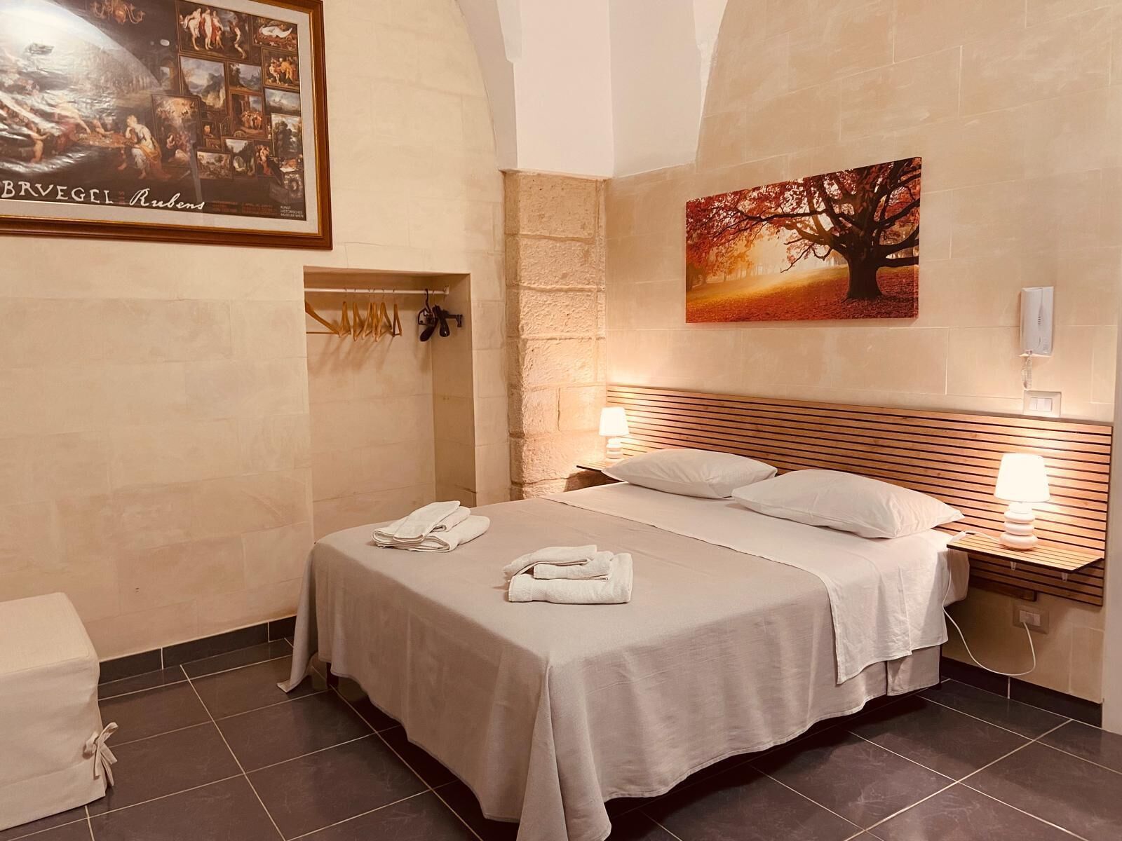 Una habitación de hotel con una cama grande, cabecera, dos mesitas de noche con lámparas, un cuadro de un árbol y un marco con una fotografía en la pared.
