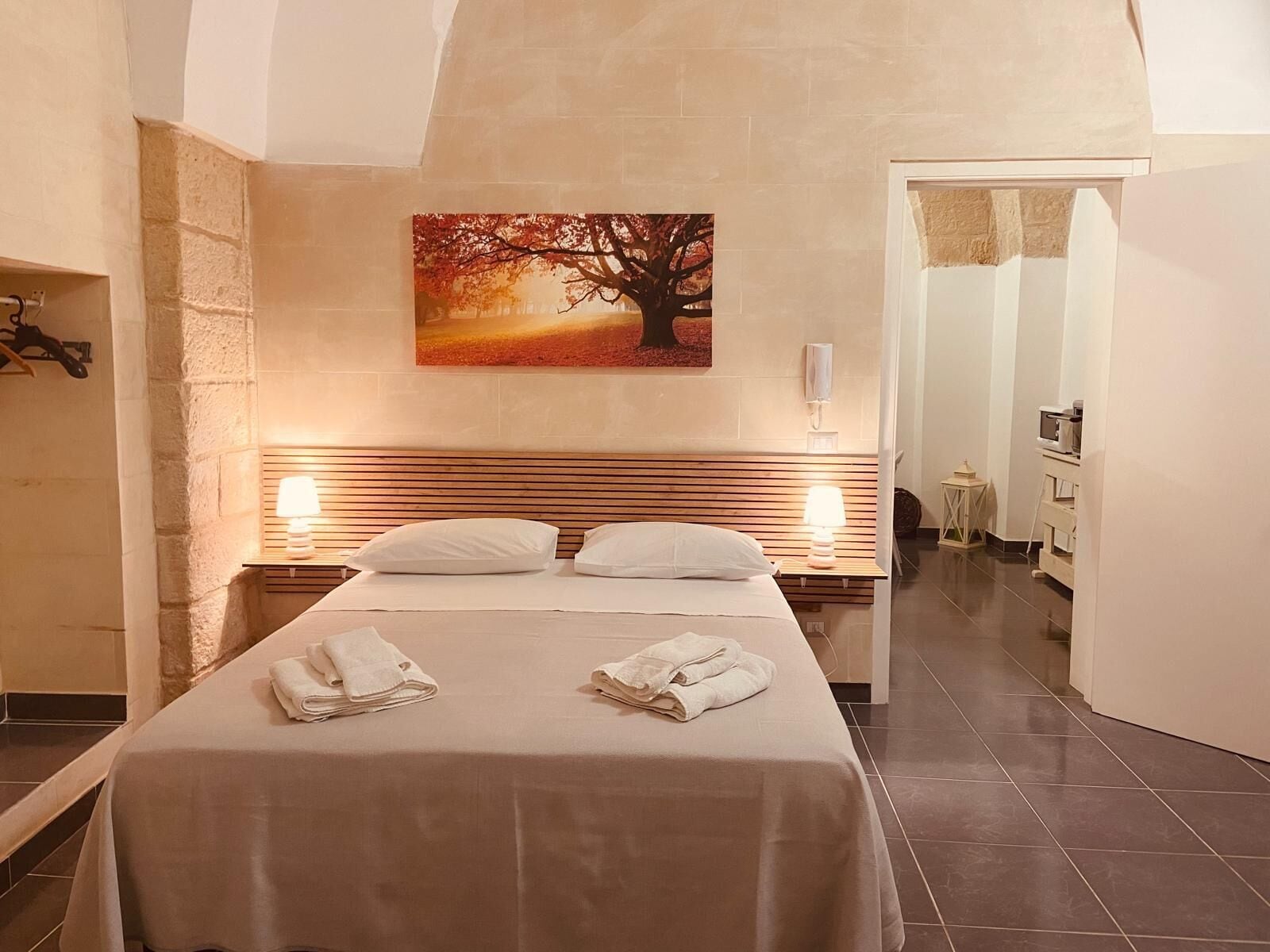 A hotel room with a bed, two bedside lamps, a painting of a tree, and a bathroom visible through an open door.