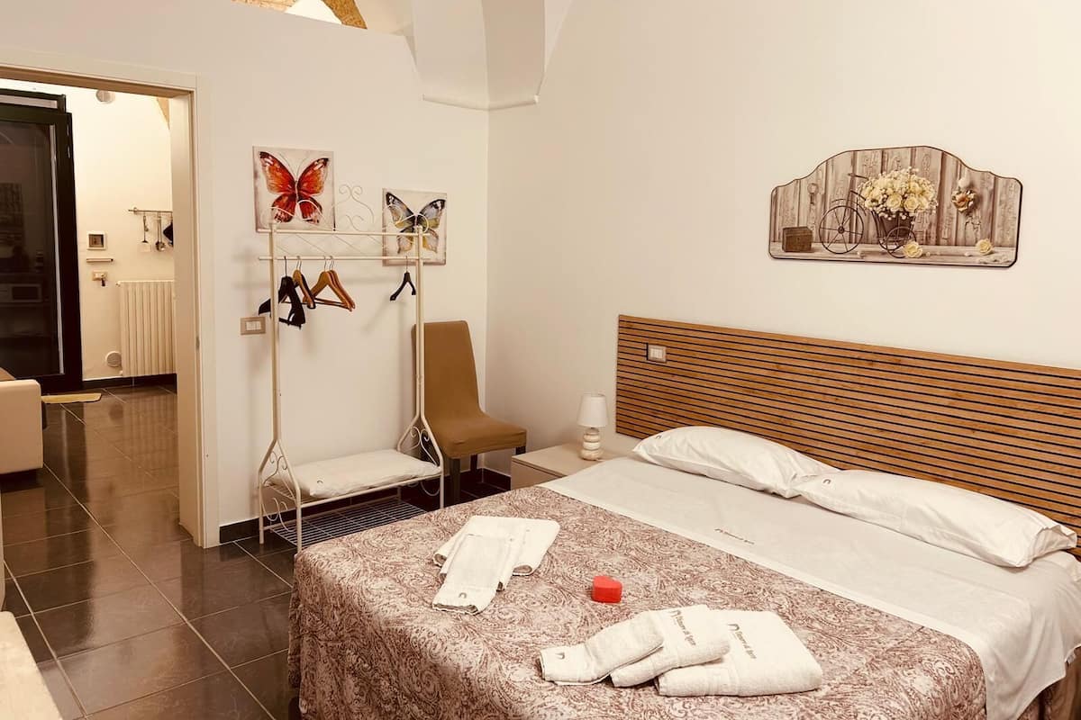 A hotel room with a bed, a chair, a coat rack with hangers, a radiator, and a window with a view of the outside.