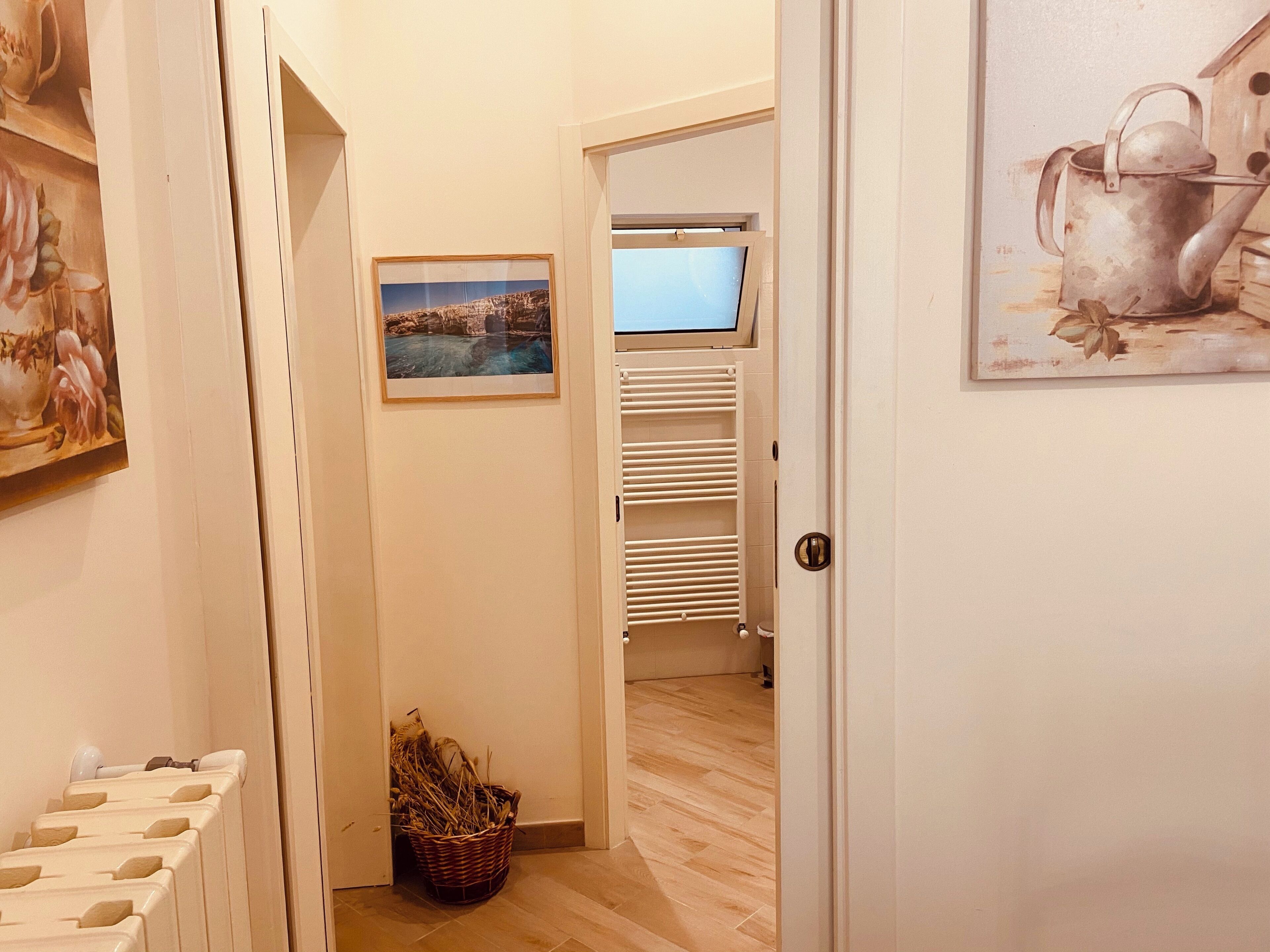 Um corredor com chão de madeira, um radiador branco, uma cesta de vime, um quadro emoldurado e uma porta branca que dá para um quarto com janela e uma estante branca.