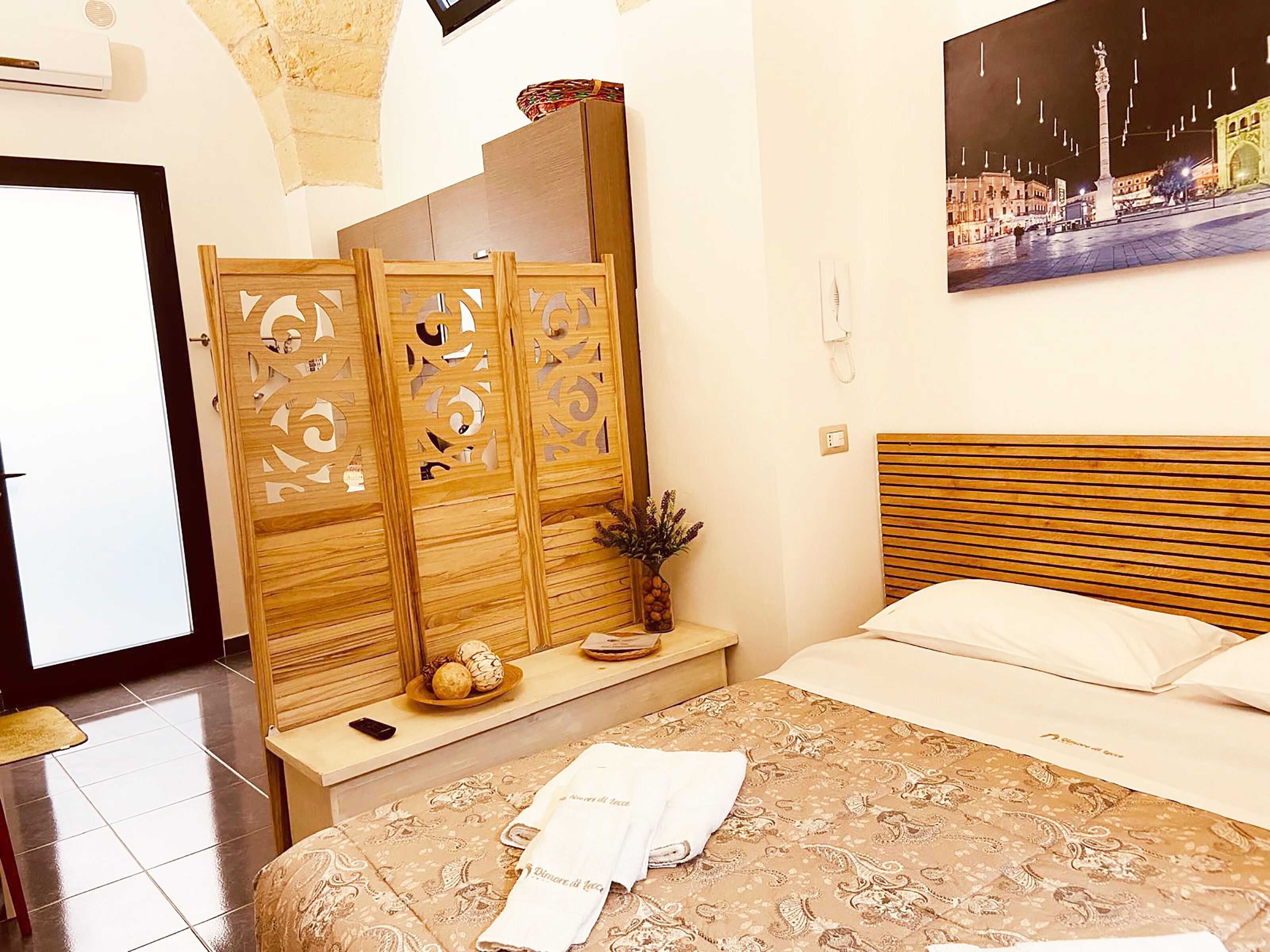 A hotel room with a bed, a wooden headboard, a decorative screen, a small table with a vase, and a painting on the wall.