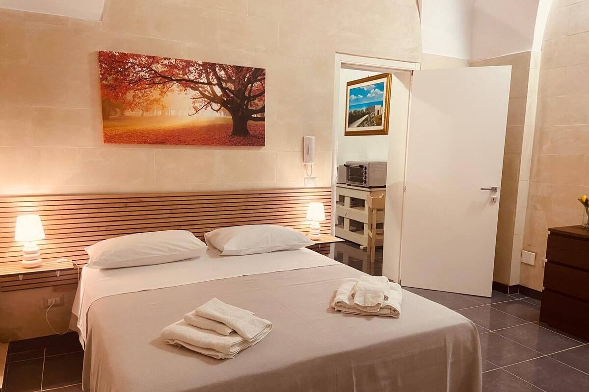 A hotel room with a bed, two bedside lamps, a painting of a tree, a door with a picture frame, and a small table with a lamp.