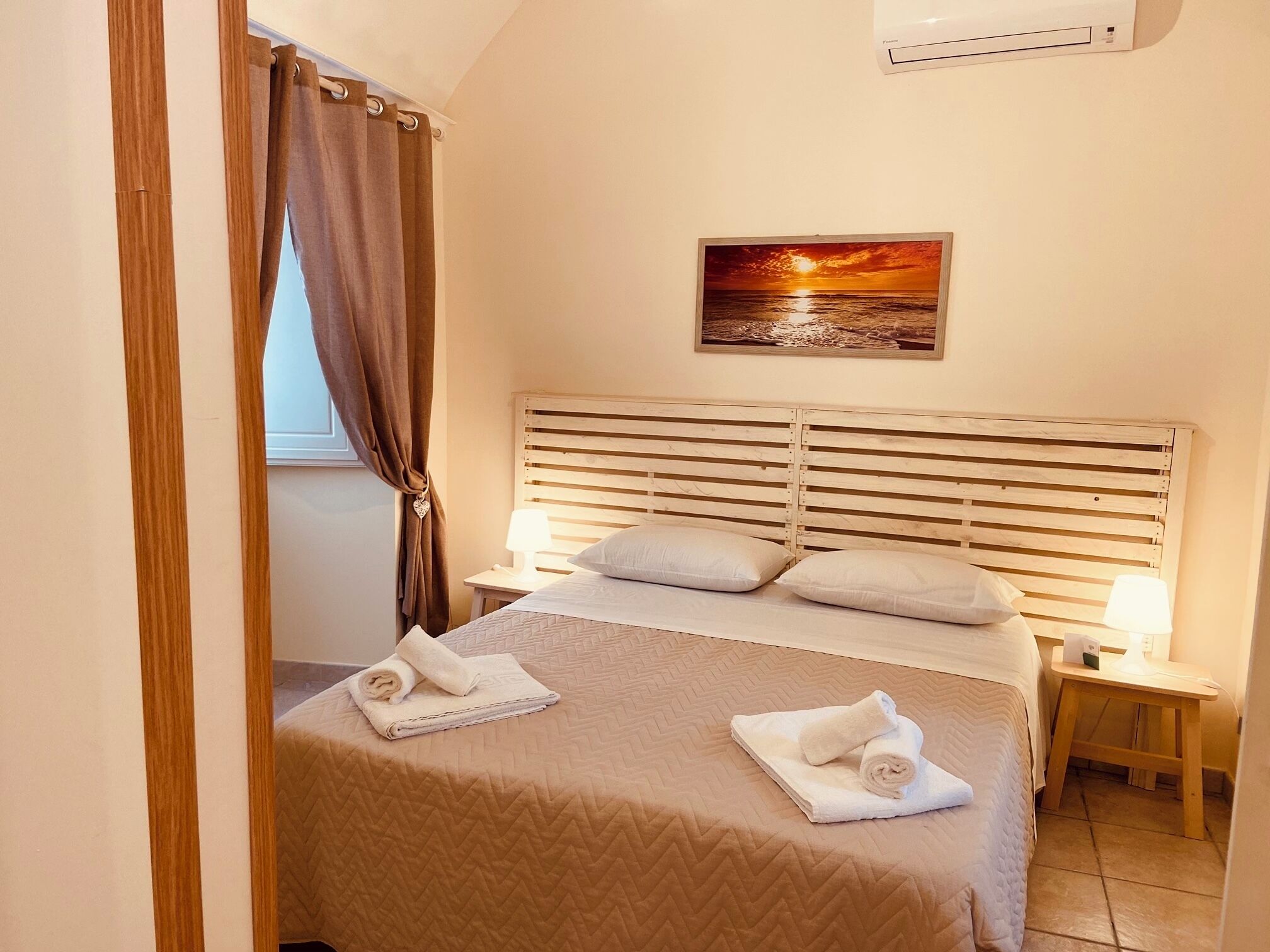 Een slaapkamer met een houten hoofdbord, twee nachtkastjes met lampen, een bed met een gewatteerde spreideken en een schilderij van een zonsondergang aan de wand.