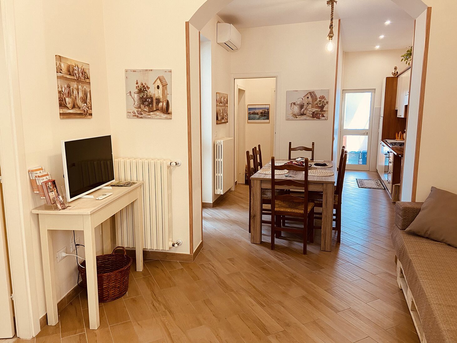 A living room with a TV, a dining table, and chairs, a kitchen in the background, and a hallway leading to another room.