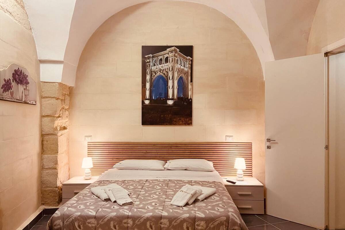 A hotel room with a large bed, a nightstand, a lamp, and a painting of a building on the wall.