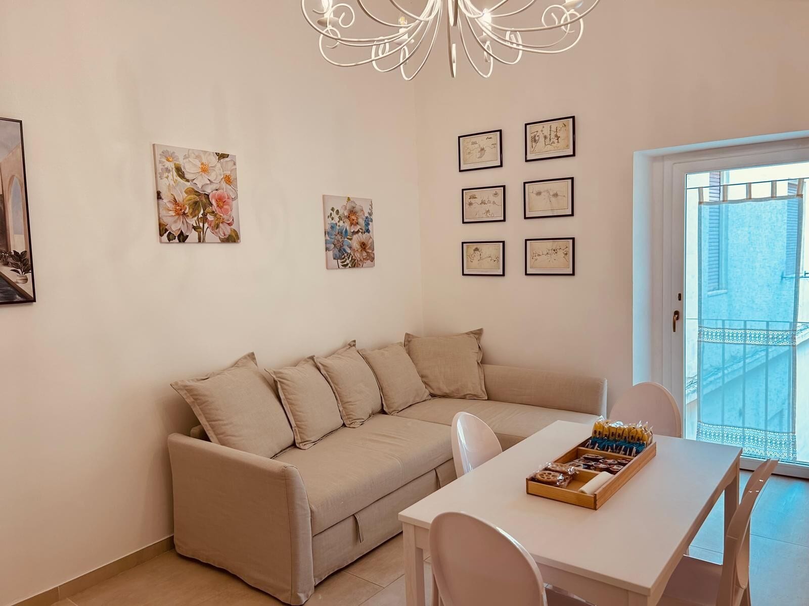 A cozy living room with a beige sofa, a white dining table, and a chandelier. There are six framed pictures on the wall.