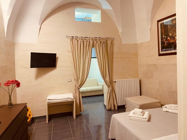 A hotel room with a bed, a small seating area, a radiator, a TV mounted on the wall, and a window with curtains. - Dimore di Lecce (Lecce)