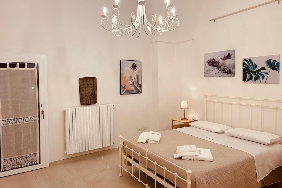 A bedroom with a bed, a radiator, a hanging towel, a chandelier, and two wall-mounted paintings.
