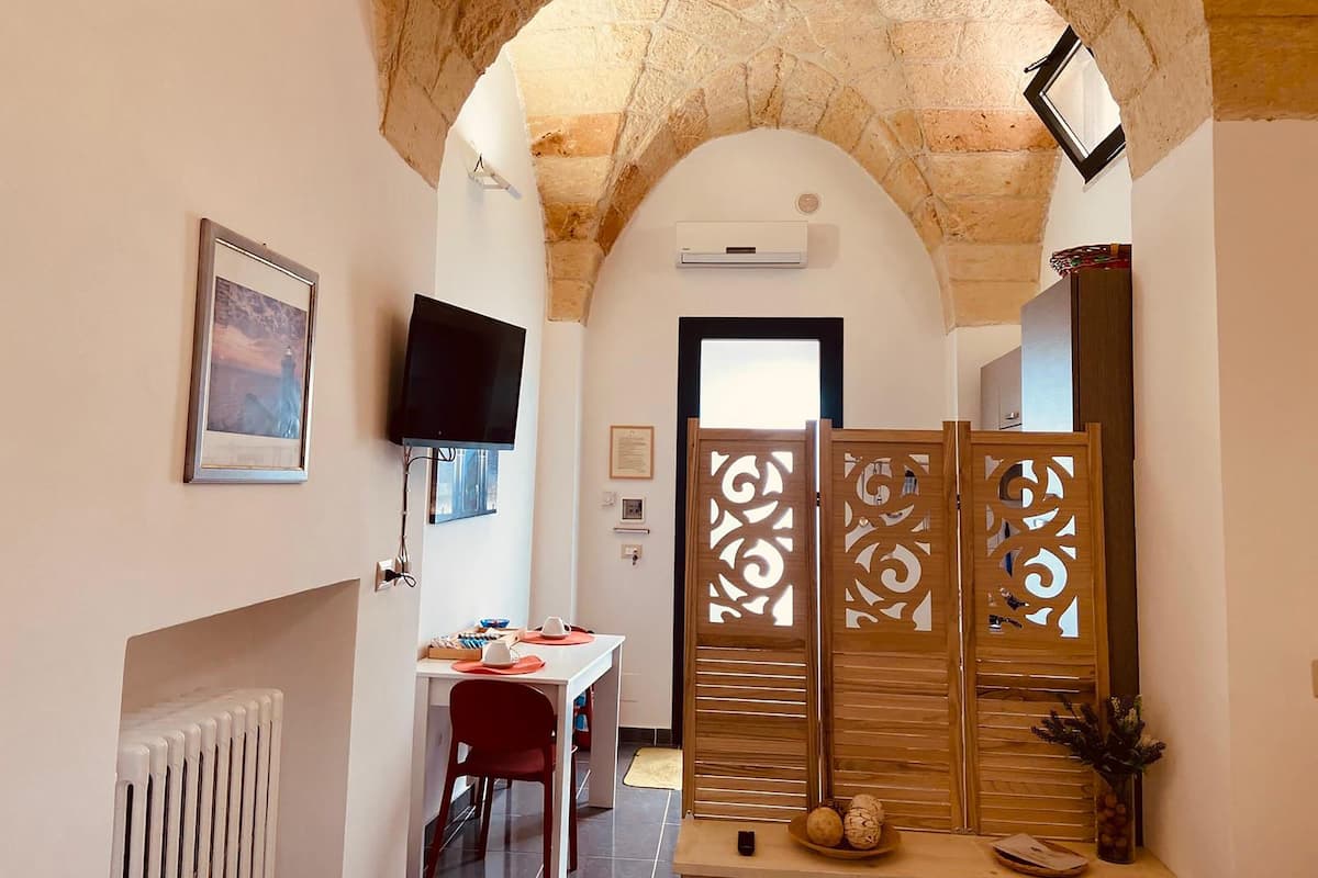 A narrow hallway with a stone ceiling, a television mounted on the wall, a white radiator, a dining table with chairs, and a decorative wooden partition.