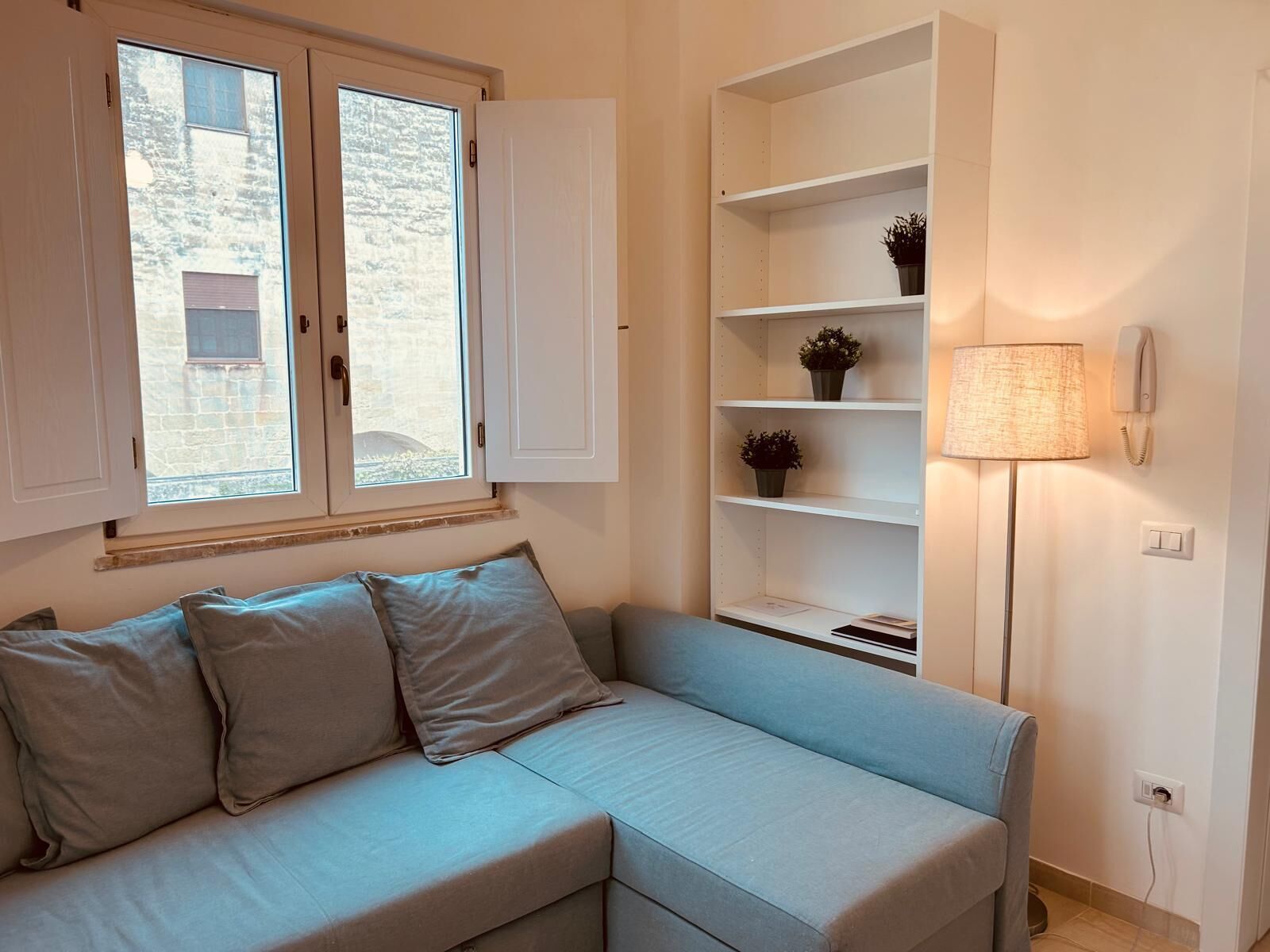Una habitación con un sofá azul claro, dos armarios blancos, una estantería con plantas en macetas, una lámpara de pie y un teléfono en la pared.