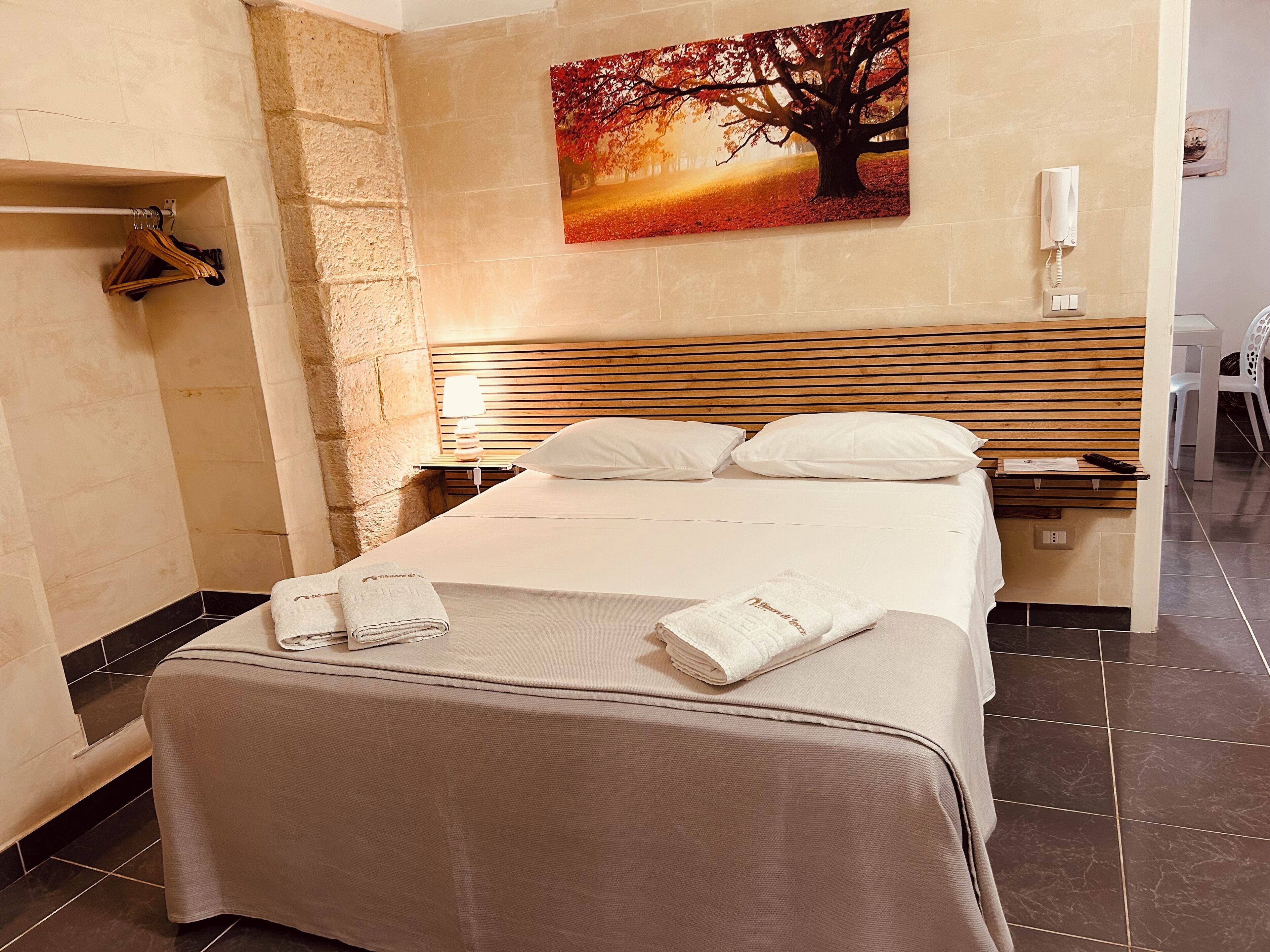 A hotel room with a bed, a wooden headboard, a nightstand, a lamp, a painting of a tree, a towel, and a chair.