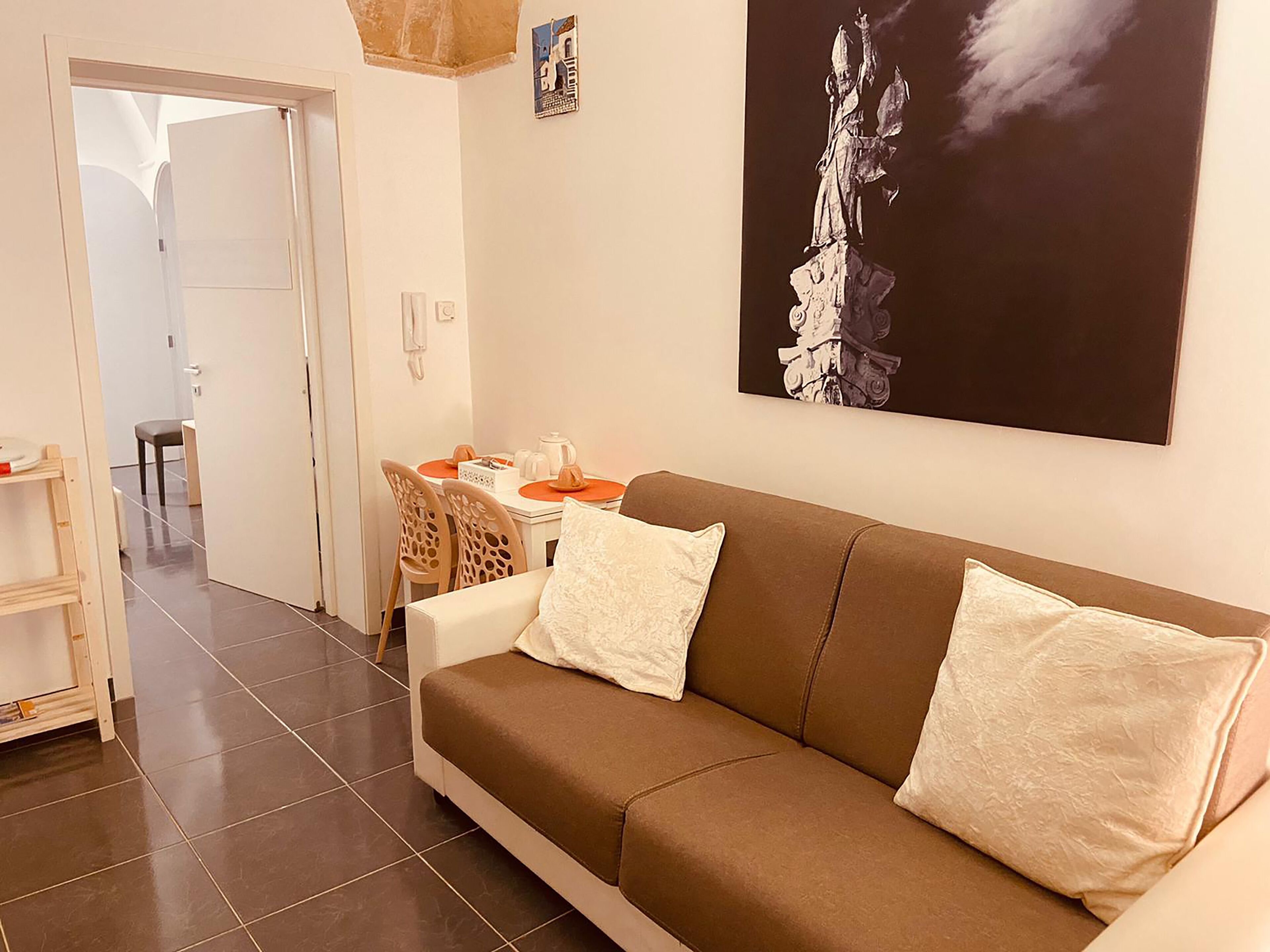 A living room with a brown sofa, two white cushions, a dining table with orange placemats, and a painting of a statue on the wall.