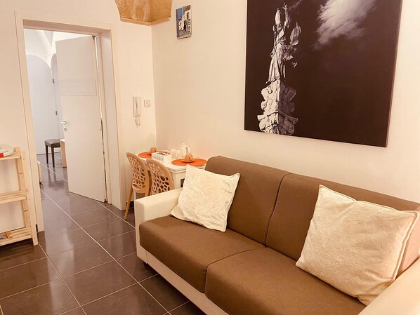 Un salón con un sofá marrón, dos cojines blancos, una mesa de comedor con manteles individuales naranja y un cuadro de una estatua en la pared.