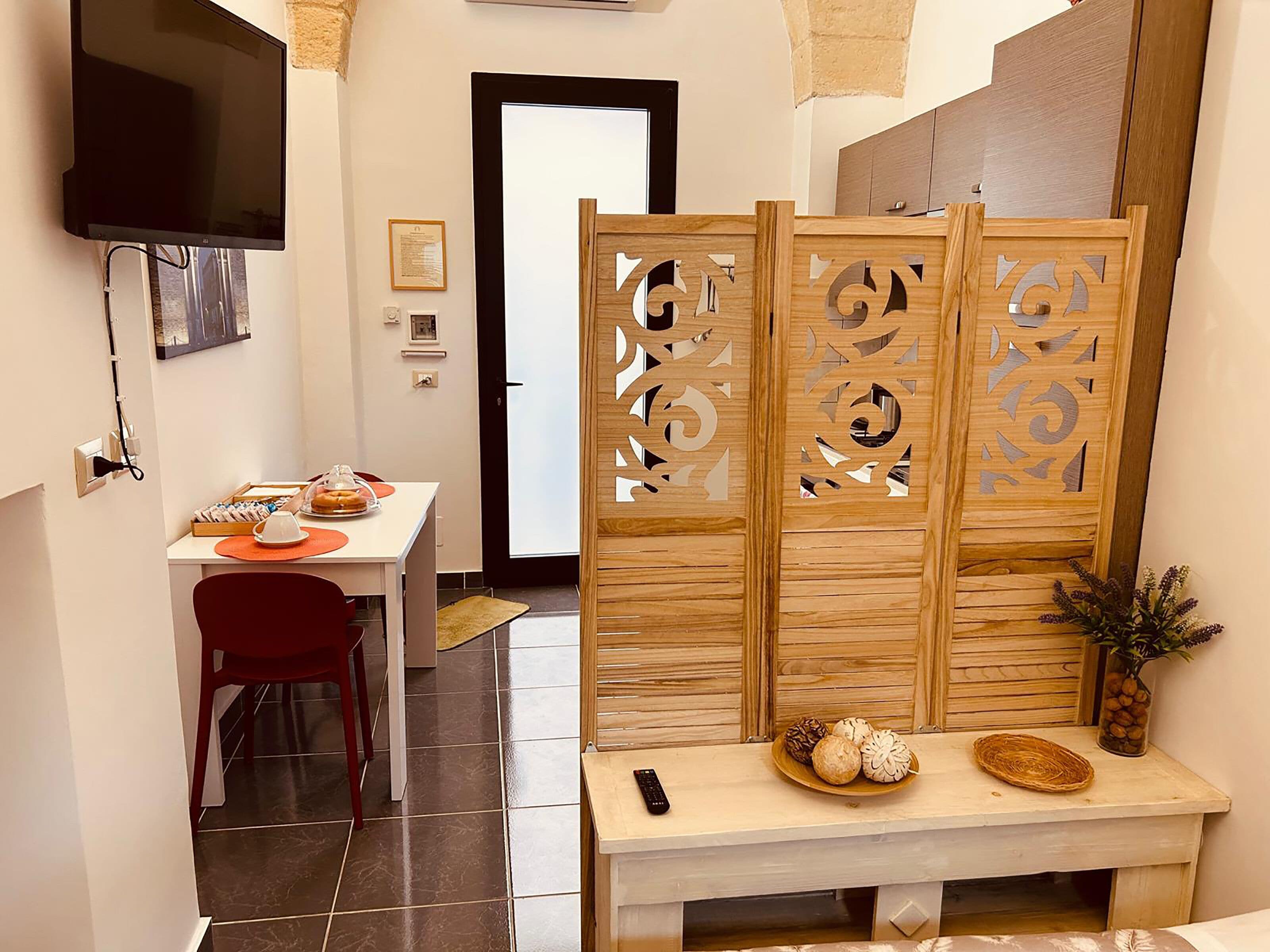A hotel room with a dining area, a TV mounted on the wall, a wooden partition with cut-out designs, a small table with a remote control, and a decorative vase with plants.