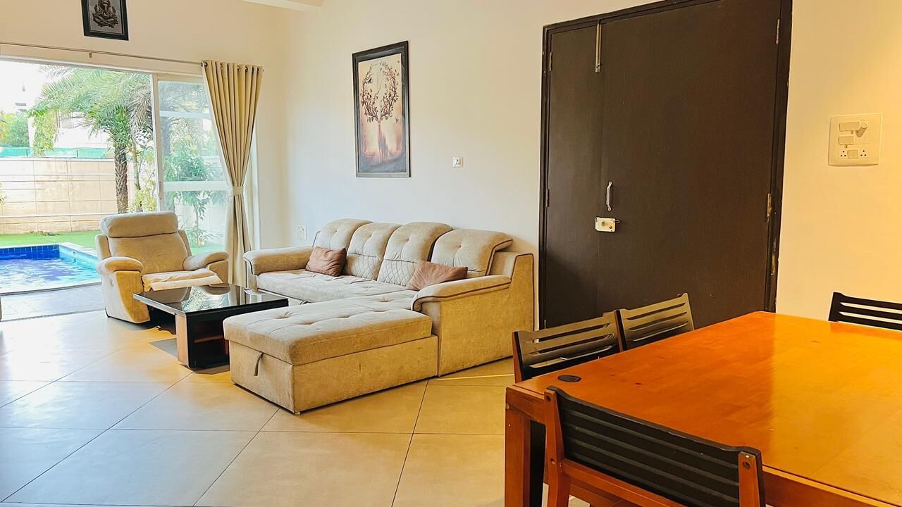 A living room with a sofa, armchair, ottoman, dining table, and chairs. There is a sliding glass door leading to a pool.