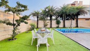 A patio with a table and chairs, a swimming pool, and palm trees. - Ashford 4 BHK With Pvt Pool By Stellar Stays (Mawal)
