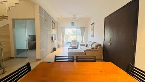 A dining area with a wooden table and chairs, a sofa, a television, and a sliding glass door leading to a balcony. - Ashford 4 BHK With Pvt Pool By Stellar Stays (Mawal)