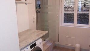 Een wasruimte met een wasmachine, een klein plankje en een raam met uitzicht op de omgeving.