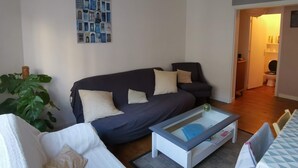 Een moderne woonkamer met een donkerblauwe bank, een glazen salontafel en een rode hanglamp. Een grote plant is in de hoek te zien, en een lijst met collages hangt aan de muur.