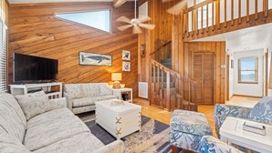 A spacious living room with a large TV, wooden walls, a staircase, and a ceiling fan. - Summer Days 4 Bedroom Oceanfront Home (Kill Devil Hills)