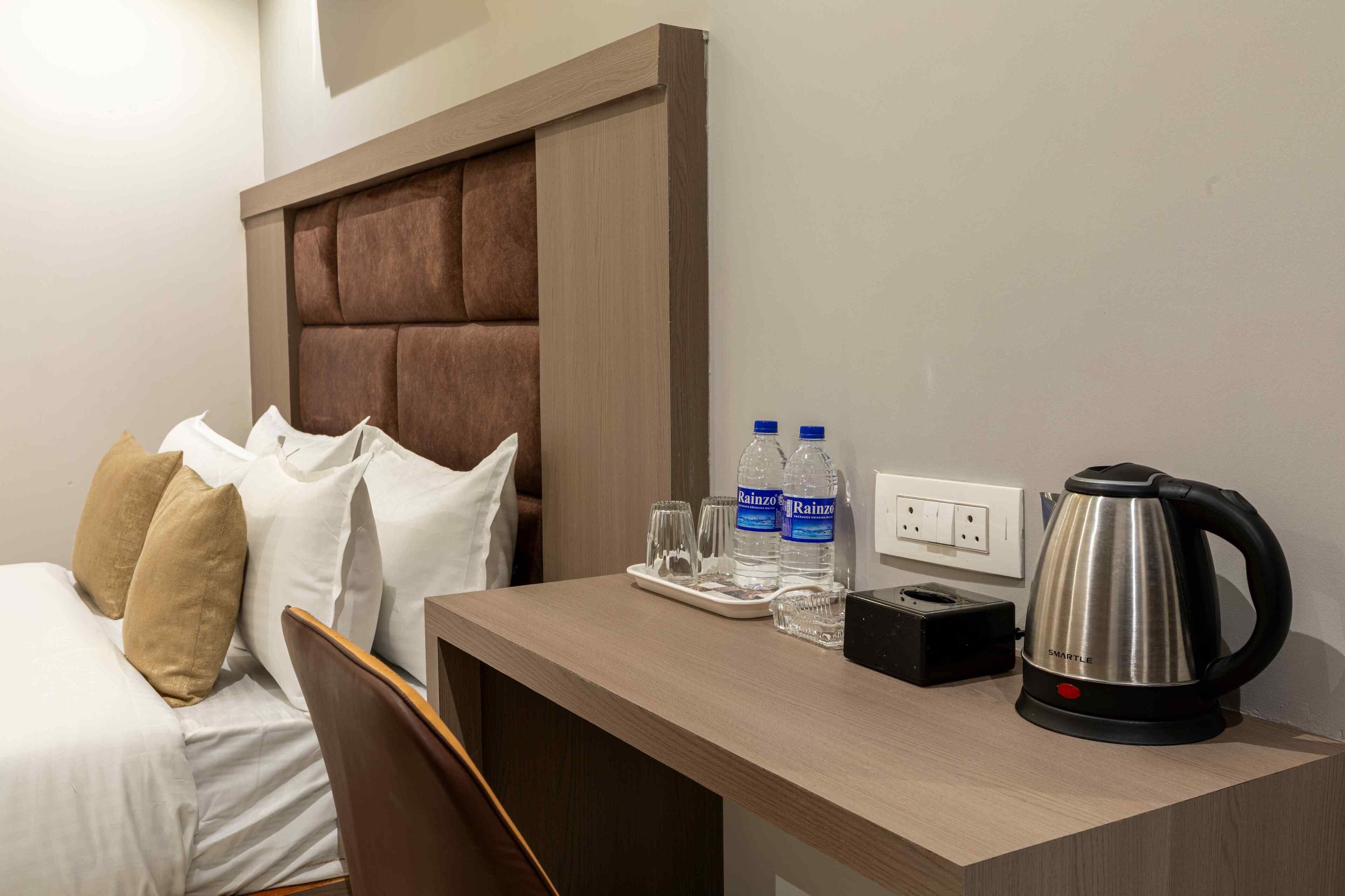 Quarto de hotel com cama, cabeceira de madeira, escrivaninha com chaleira, copos e garrafas de água.