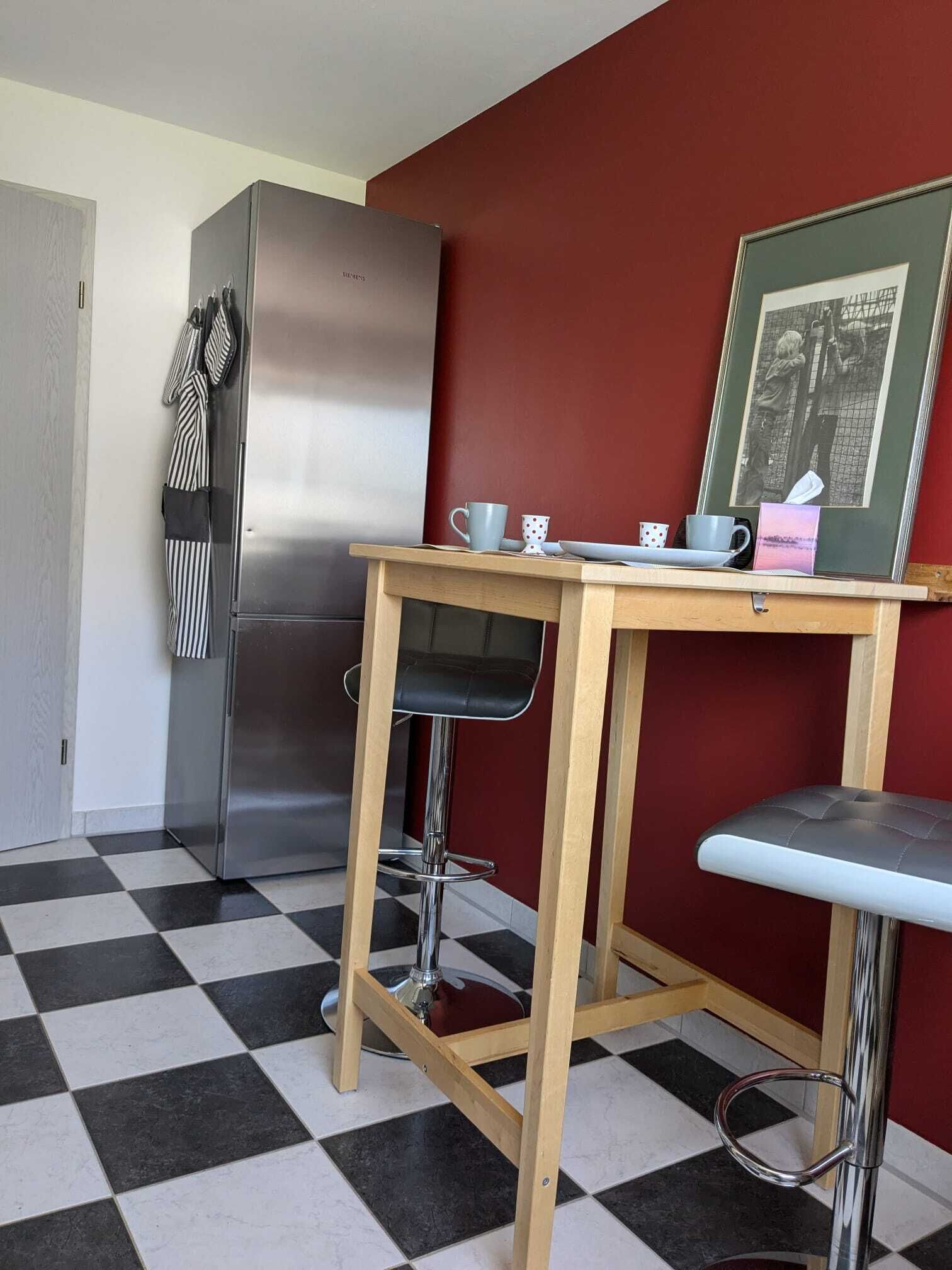 Eine kleine Küche mit einem Edelstahl-Kühlschrank, einem Holztisch mit zwei Barhocker und einem gerahmten Bild an der Wand.