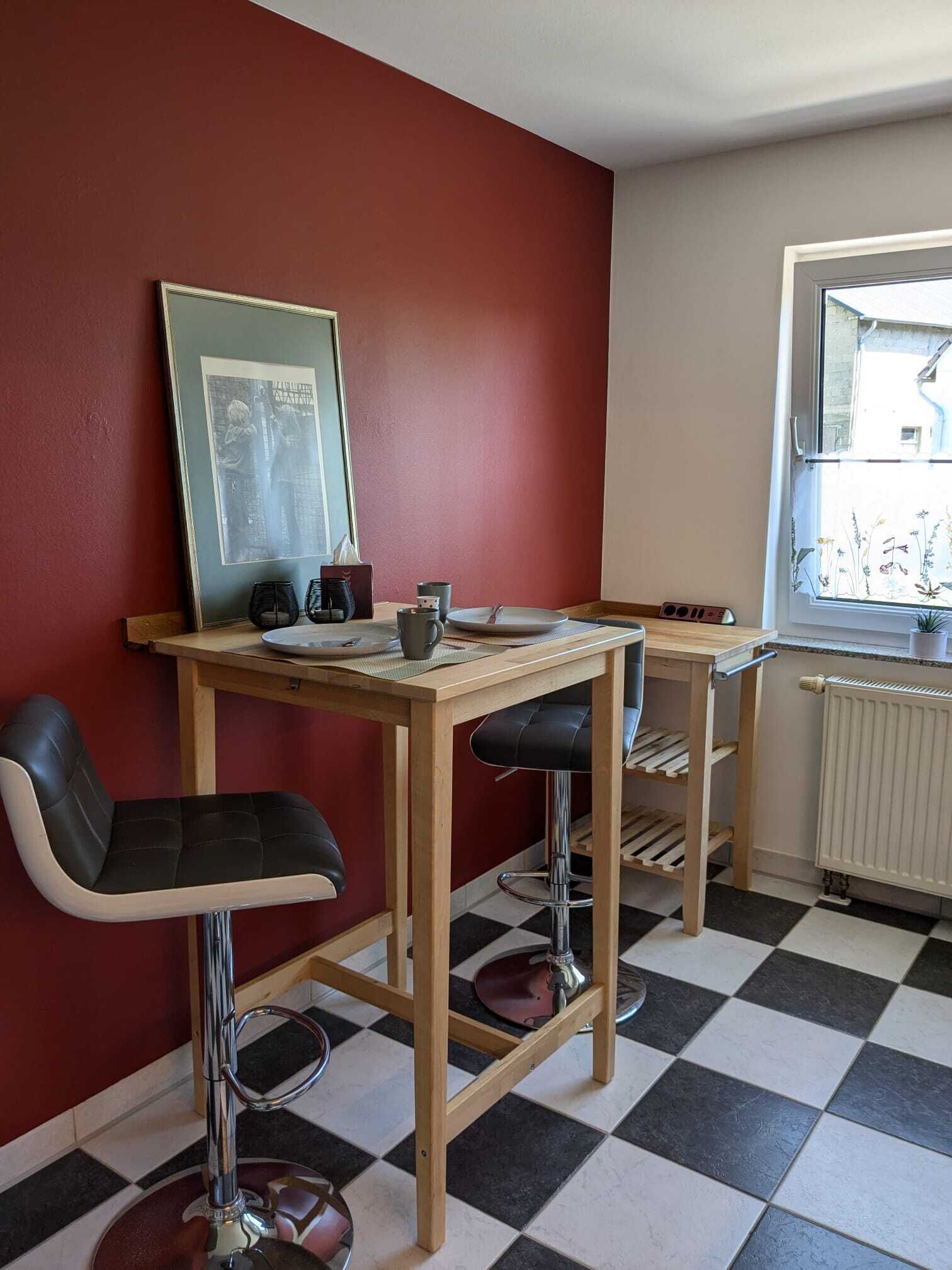 En matplats med ett rutmönstrat golv, ett träbord, två svarta barstolar, en inramad tavla på väggen och ett fönster med utsikt över byggnader.