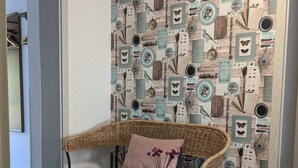 Une chaise en osier avec un coussin rose et un vase rempli de fleurs violettes, placée devant un mur recouvert d’un papier peint au motif rétro.