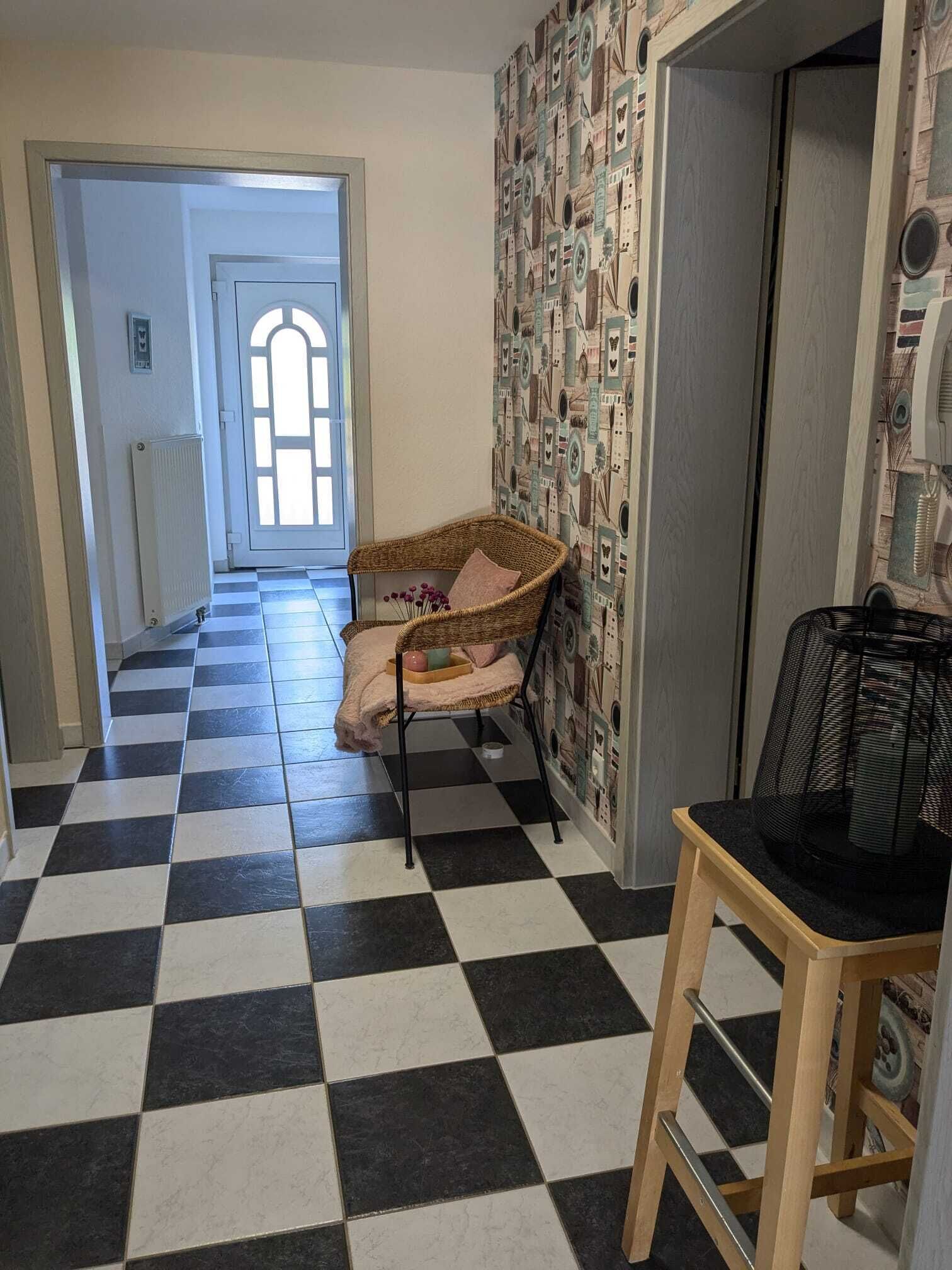 A hallway with a checkered floor, a wicker chair with cushions, a radiator, and a patterned wallpaper.