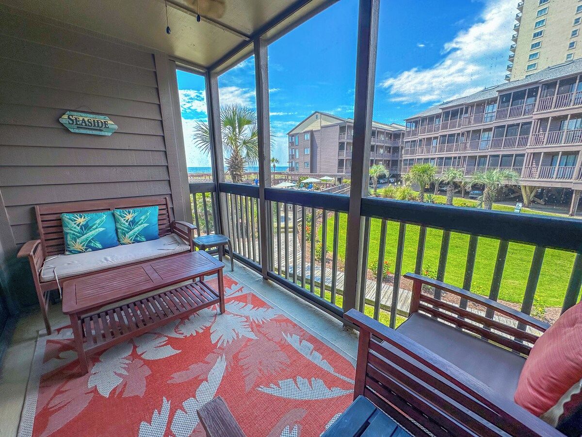 A balcony with a wooden bench and a small table, a red patterned rug, and a view of a hotel building and a beach.