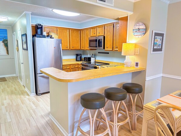 A compact kitchen with wooden cabinets, a stainless steel refrigerator, a microwave, and a coffee maker. There are four stools at the bar counter. - Tilghman Beach and Racquet Club Unit: 117! 3 Bedroom Ocean View Condo. Pools, Tennis, Close to Main Street! (North Myrtle Beach)