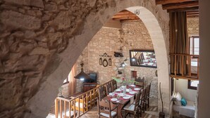 A rustic stone interior with a wooden dining table set for four, a television, and a cozy seating area with cushions. - Kritiko Archontiko, Nature & Private Pool Escape

 (C6Q2+MP Aspro, Greece)