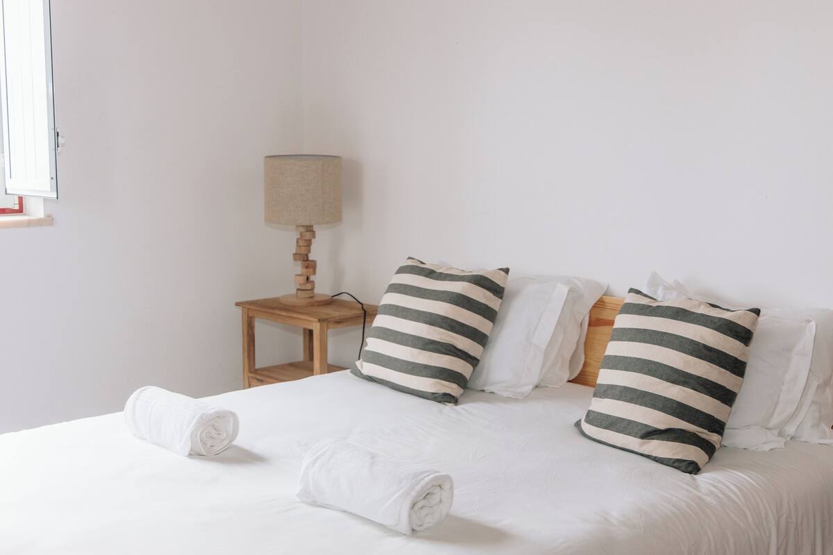 A neatly made bed with striped pillows, a small wooden nightstand with a lamp, and a rolled towel on the bed.