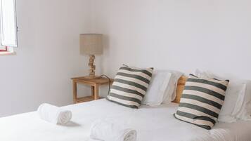 Una cama bien hecha con almohadones a rayas, una mesita de madera pequeña con una lámpara, y una toalla enrollada sobre la cama.