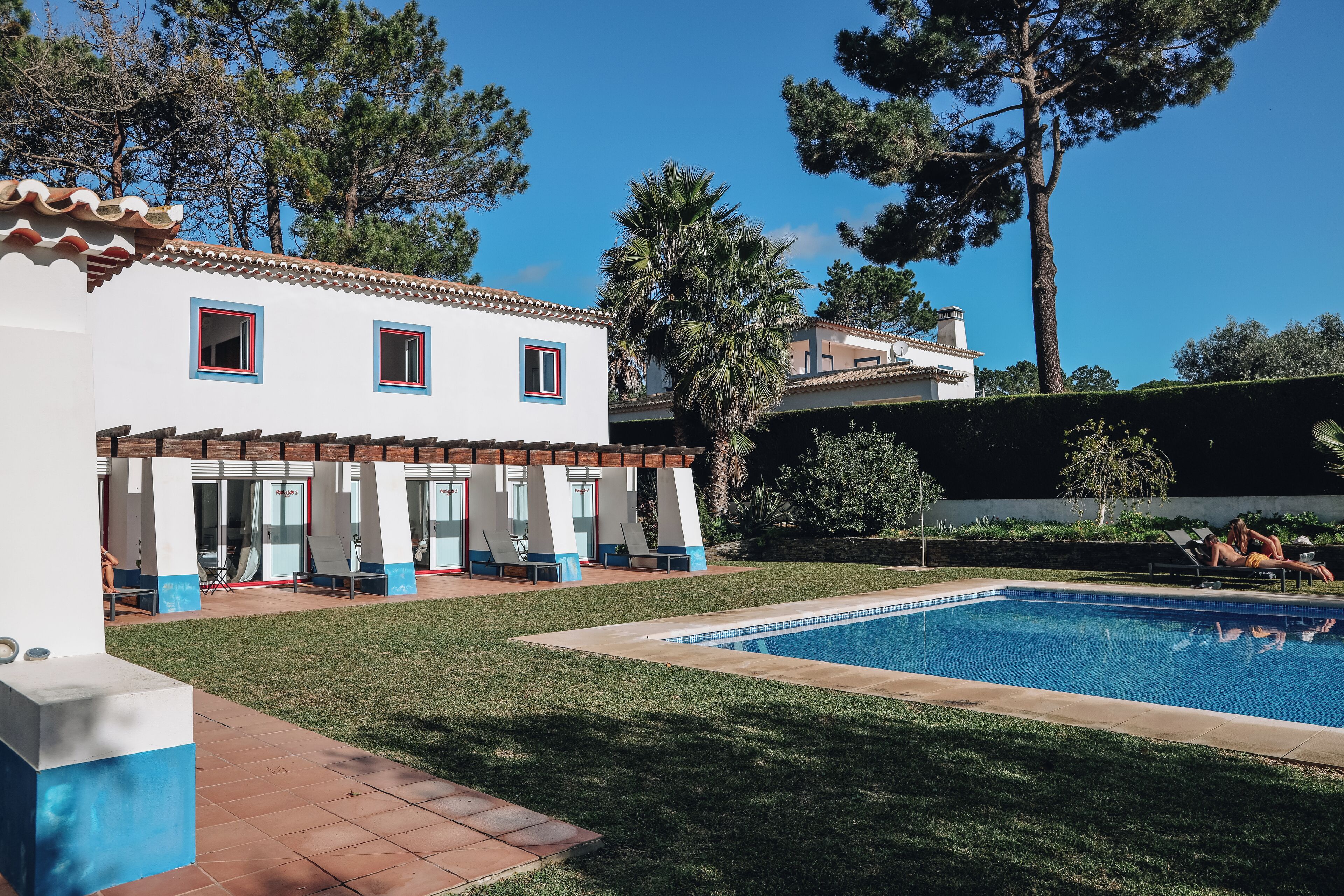 A pool area with a clear blue pool, a white building with red window frames, and a well-maintained lawn.