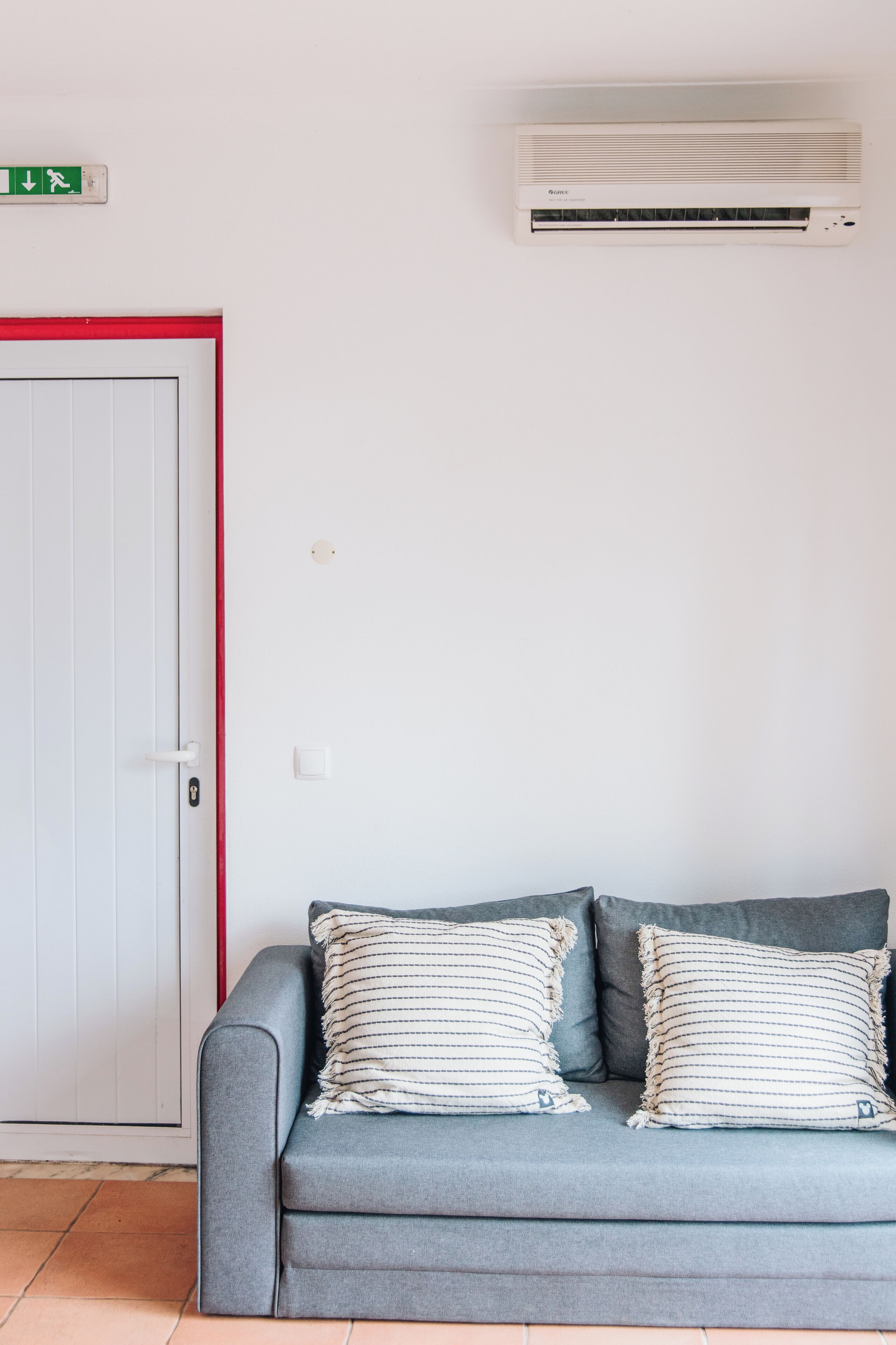 A room with a grey sofa, two striped pillows, a white door with a red frame, and a wall-mounted air conditioner.