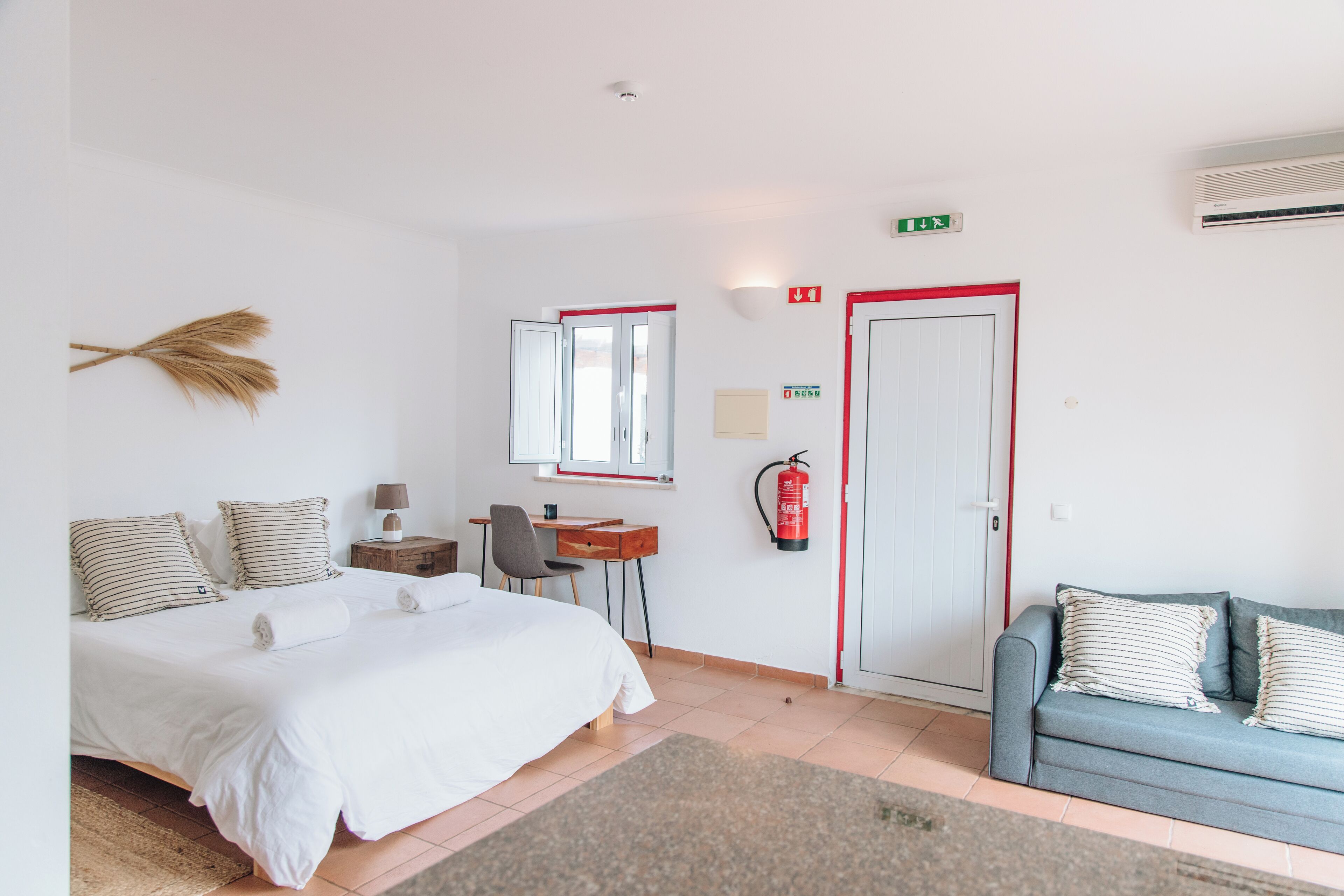 A hotel room with a bed, a desk, a chair, a sofa, a mirror, a fire extinguisher, and an exit sign.