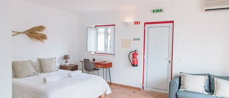 A hotel room with a bed, a desk, a chair, a sofa, a mirror, a fire extinguisher, and an exit sign.
