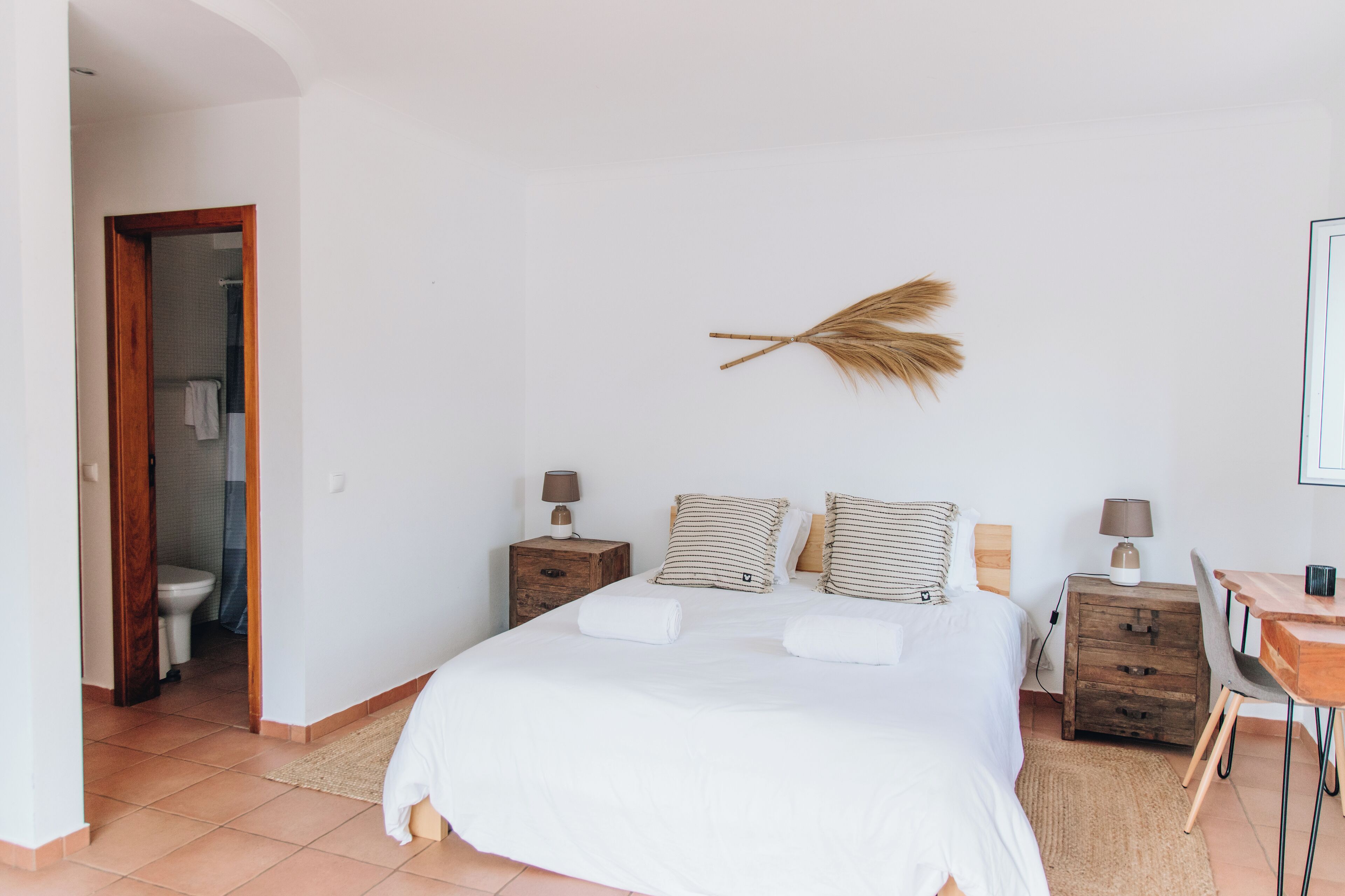 Un dormitorio con una cama grande, dos mesitas de noche con lámparas, una mesita de noche de madera, un escritorio con una silla y una pluma decorativa en la pared.