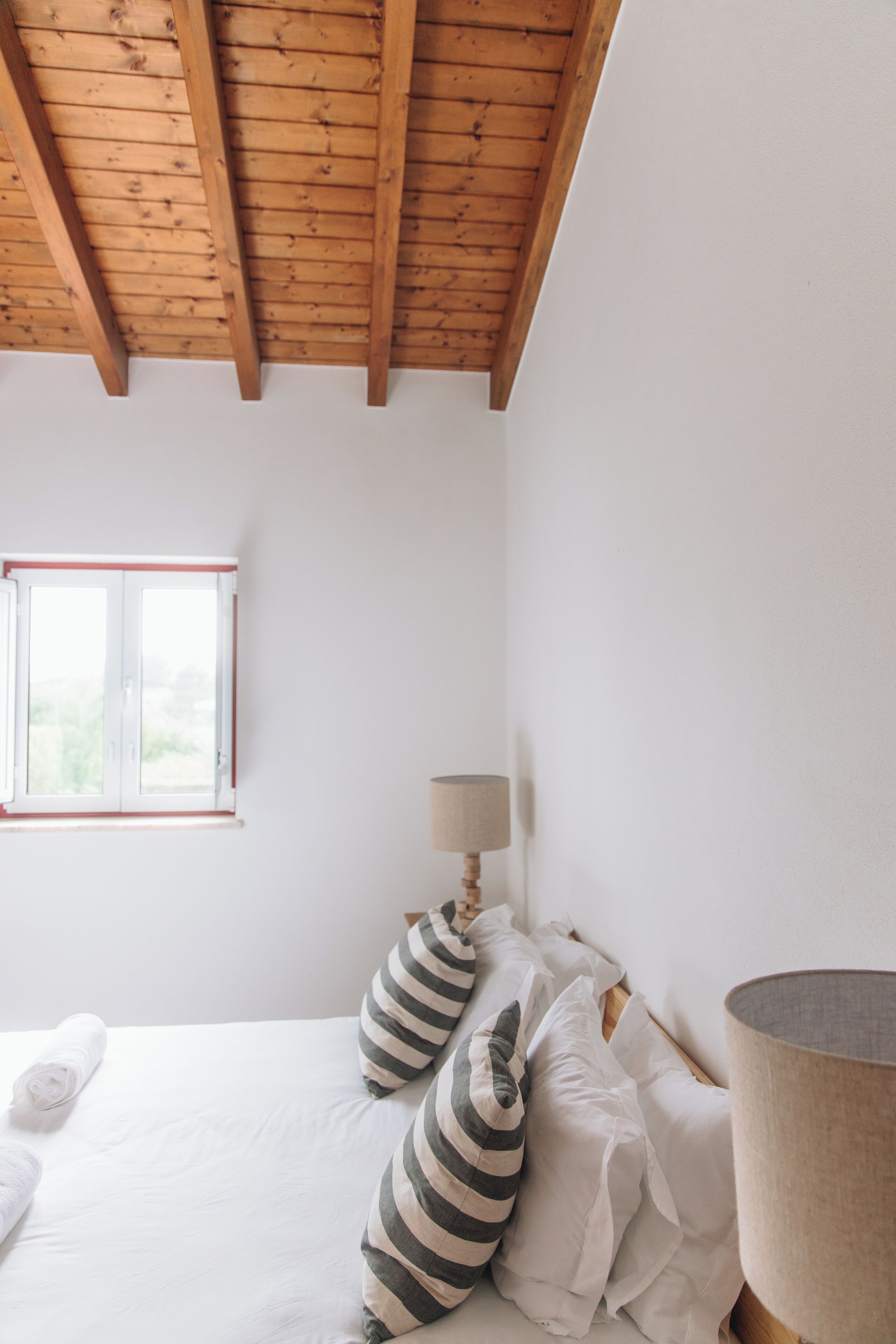 A bedroom with a wooden ceiling, a window, a bed with white bedding and striped pillows, and a bedside lamp.