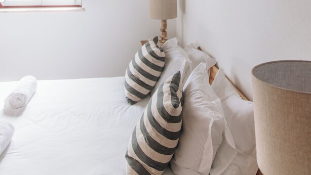 Een slaapkamer met een houten plafond, een raam, een bed met witte lakens en gestreepte kussens, en een bedlampje.