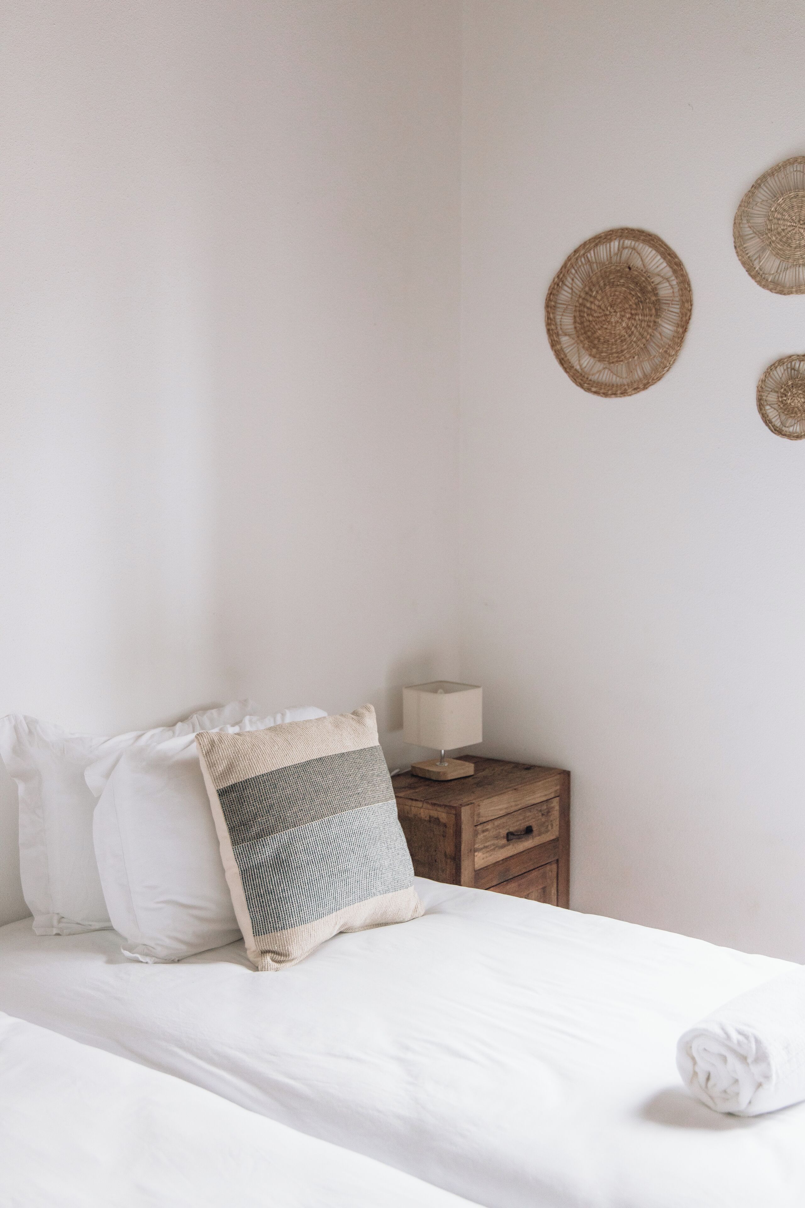 A neatly made bed with white bedding and a grey and white striped pillow. A wooden nightstand with a lamp and a small drawer is beside the bed.