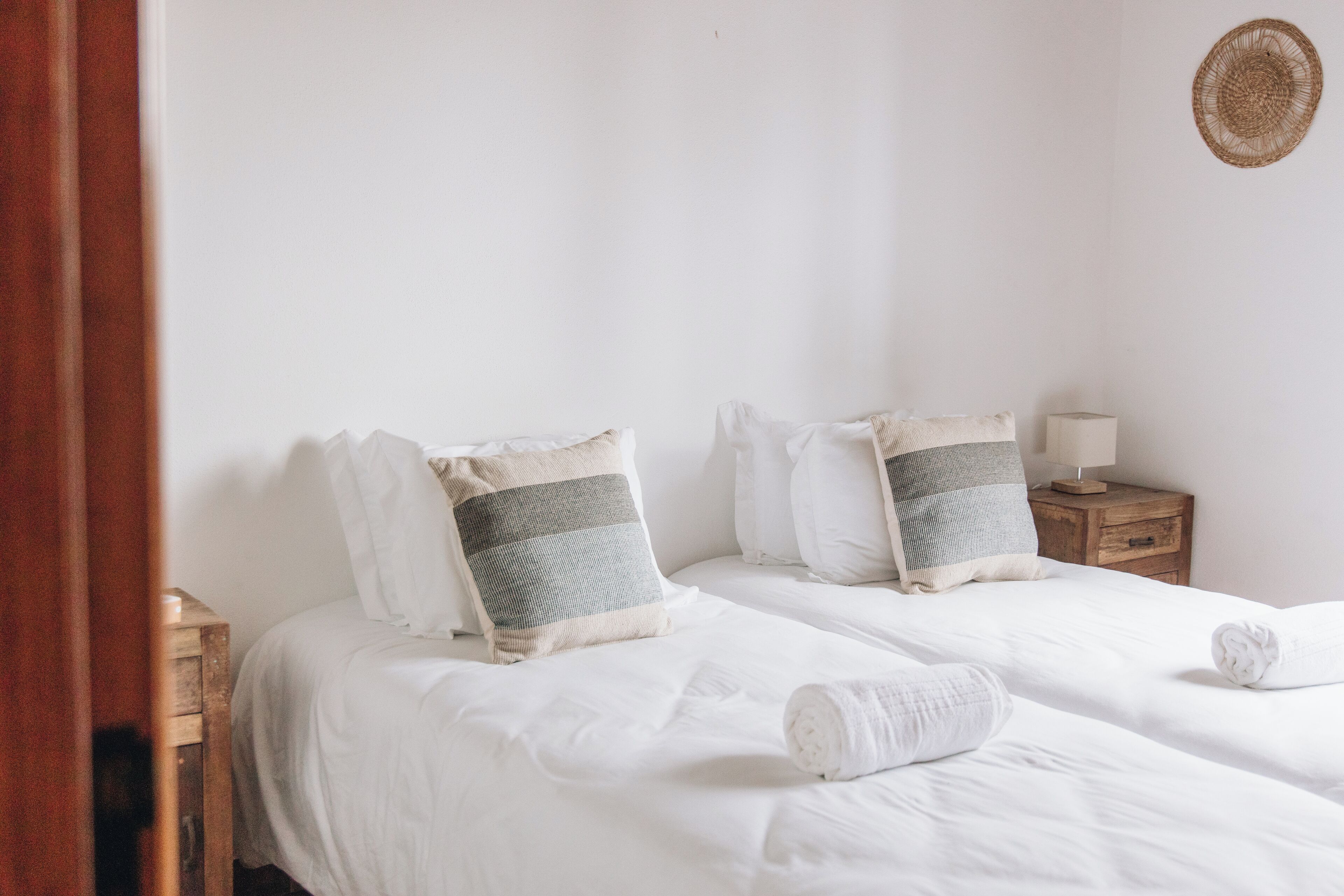 A neatly made twin bed with white linens and striped pillows, a wooden nightstand with a lamp, and a woven wall hanging.