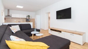 Ein stilvolles Wohnzimmer mit einer Couch, einem kleinen Tisch, einem an der Wand befestigten Fernseher und einer Küche im Hintergrund.