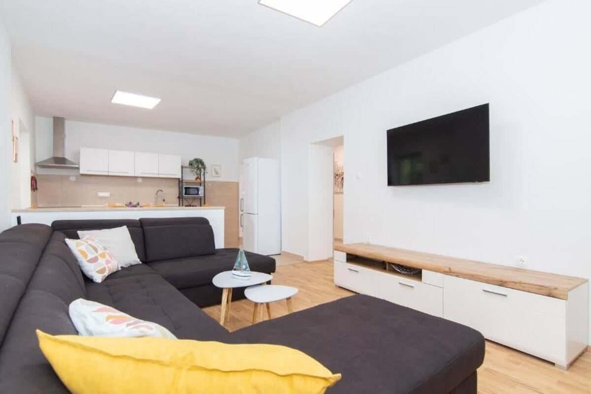 Ein stilvolles Wohnzimmer mit einer Couch, einem kleinen Tisch, einem an der Wand befestigten Fernseher und einer Küche im Hintergrund.