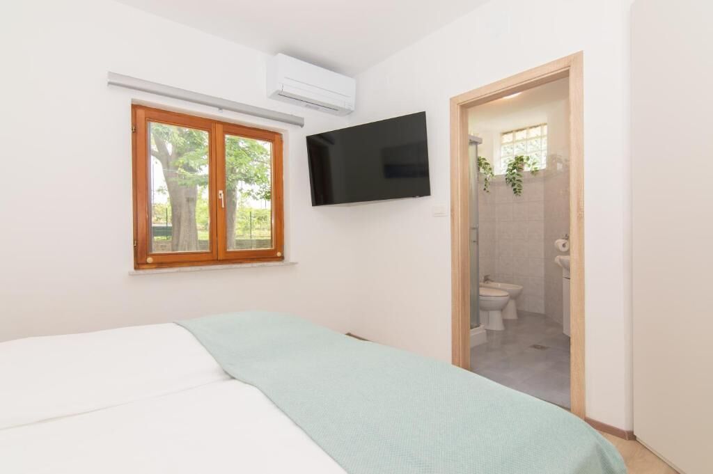 A bedroom with a bed, a TV mounted on the wall, and a window with a view of trees.