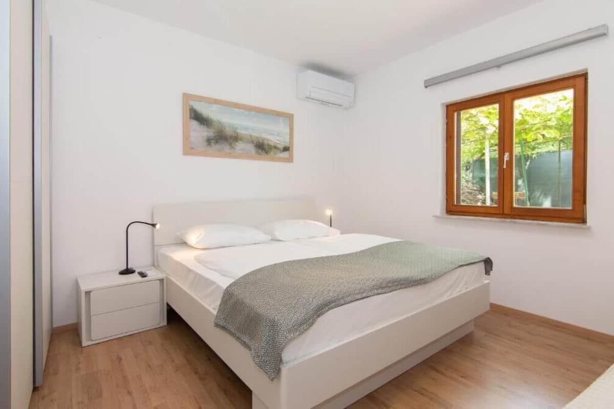 Ein Schlafzimmer mit einem Bett, einem Nachttisch, einem Gemälde und einem Fenster mit Blick auf Grünflächen.