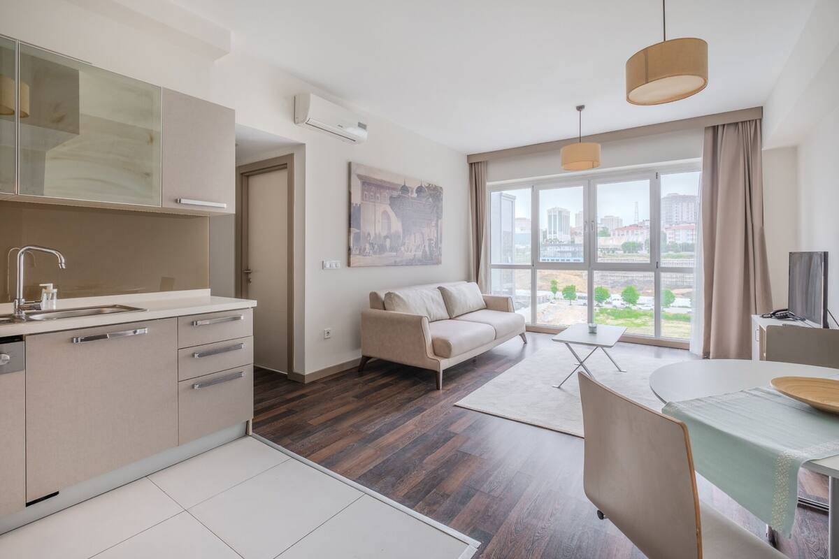 A modern apartment with a kitchen, a living area with a sofa and a dining table, and large windows offering a city view.