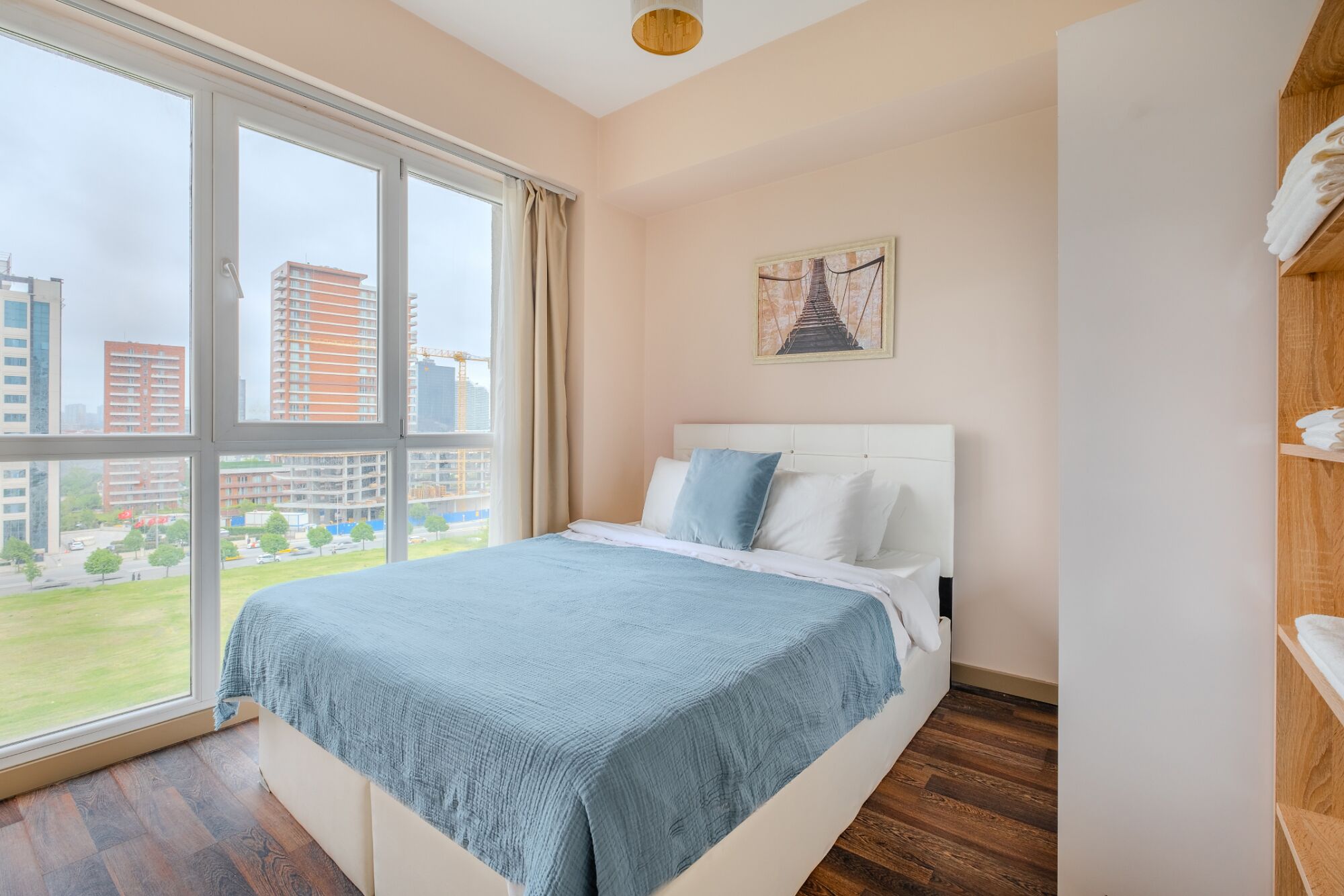 A bedroom with a large window, a bed with a blue blanket, white pillows, and a wooden floor.